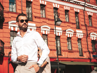 Fototapeta premium Portrait of handsome confident stylish hipster lambersexual model.Sexy modern man dressed in white shirt and trousers. Fashion male posing on street background in Europe city at sunset. In sunglasses