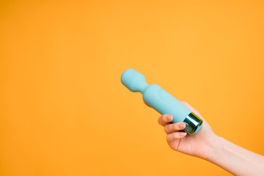 A woman's hand holds a blue dildo or vibrator for sex on a yellow background.Useful for a sex shop