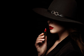 Beauty portrait of elegant woman with luxury lips makeup holding red lipstick. Model hides her face under wide hat, posing in studio, on black background. Close up. Copy, empty space for text