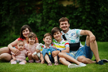 Naklejka na ściany i meble Beautiful large family with young parents and four kids sitting in green grass at courtyard.