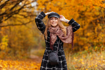 Happy beautiful young girl with a beauty smile in fashion clothes and a coat with a scarf and a hat walks in the autumn forest with yellow fall foliage