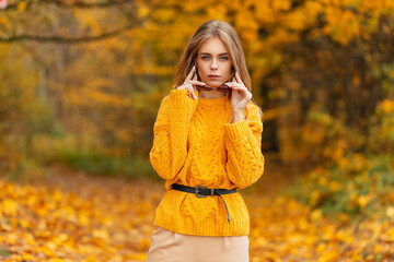 Trendy beautiful fashion young woman in knitted vintage yellow sweater dresses sunglasses outdoors on golden autumn foliage background