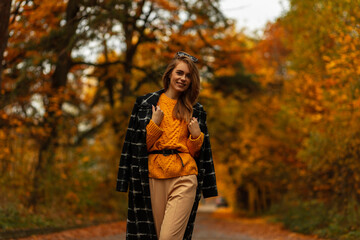 Fototapeta na wymiar Happy stylish young girl with pretty smile in vintage autumn collection clothes with knitted sweater, black coat walks outdoors with orange leaves