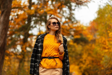 Fototapeta na wymiar Fashionable elegant beautiful girl with stylish sunglasses in vintage black coat and yellow knitted sweater in a park with autumn colored foliage