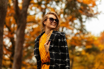 Happy woman with a cute smile with teeth in fashionable autumn clothes with a knitted sweater and a coat in nature on the background of a tree with yellow autumn foliage. Smiling female pretty face