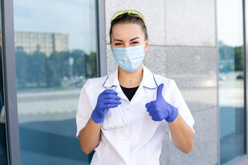 Scienсe, chemistry, biology and medicine concept - European dark-haired female scientist in a medical gown, protective mask and rubber gloves holding an Erlenmeyer flask and showing thumbs up outside.