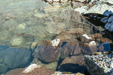 Beautiful nature background of stony bottom in transparent water of glacial lake in sunlight. Sunny nature backdrop with many stones in clear water of glacier lake. Invisible water of mountain lake.