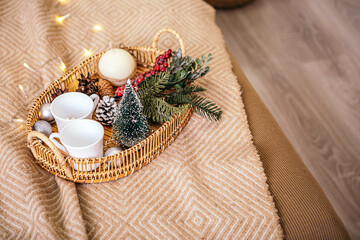 Christmas beautiful decoration for interior New Year wicker wooden tray with toys white cups cones and green branch on a warm blanket, copy space