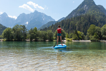 Pre-teenage boy in a protective vest standing up on the SUP board that floating on a calm lake, and use a paddle to propel through the water