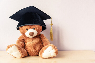 Adorable teddy bear with black academic cap sits on wooden table near light pink wall as space for...