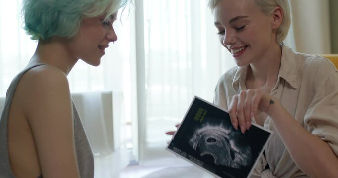 Cute Lesbian Couple Have Fun, Getting News ABout Pregnancy, Future Mothers, Side View On Happy Ladies In Casual Clothes Sit On Floor At Home, Laughing, Talking, Hugging, Celebrating Pregnancy