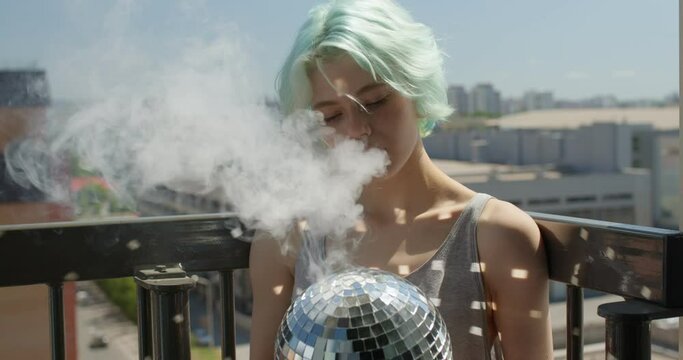 Awesome young caucasian woman with short green hair smoking electronic cigarette on balcony, holding disco ball in hands, having reflections from sun. copy space. people lifestyle, youth concept
