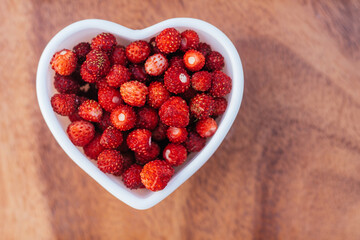 Heart shaped bowl full of ripe red juicy wild strawberries on wooden background. Top view, copy space