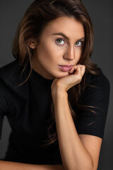 Portrait of a sexy young white woman with wavy hair and beautiful makeup sitting by herself on a table inside a studio with a grey background wearing a black shirt.