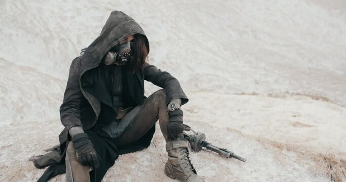 Handheld slowmotion post apocalyptic woman wearing grunge clothes hooded black mantle protective gas mask respiratory sitting on white hill in desert. Cyberpunk warrior, future female survivor
