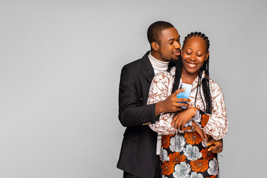 Smiling Young Black Man Embracing Woman From Behind While Looking At Smartphone. African American Couple Sharing Social Media On Mobile Phone, Happy Female