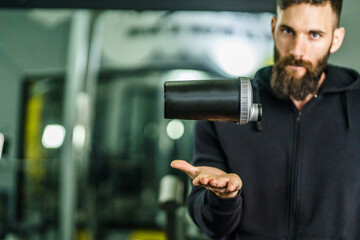 Front view portrait of young caucasian man athlete in black hoodie male standing in the gym holding protein supplement shaker supplementation in training waist up black hair and beard copy space