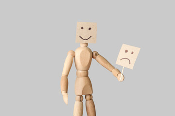 Wooden mannequin hiding face behind emoticon on light background