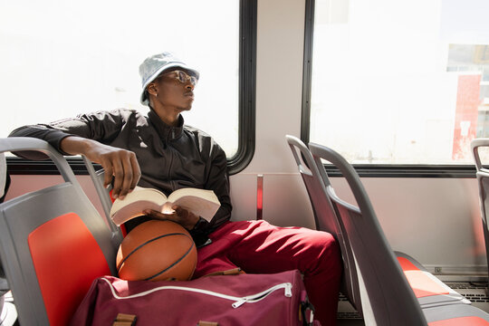 Young male college student with book and basketball on sunny bus