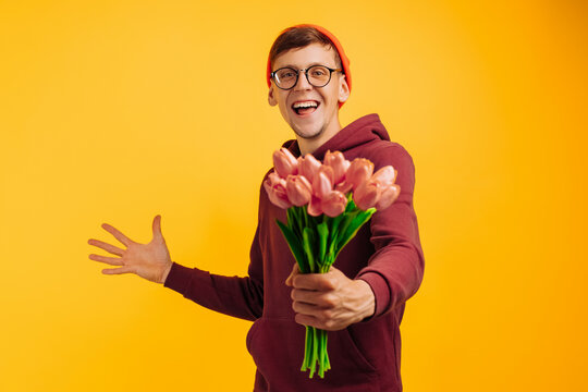 A Handsome Man With A Bouquet Of Flowers With Delight Gives Flowers Wrapped In Pink Paper, A Man In An Orange Hat