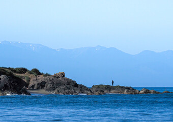 Lone person standing on rocky shore overlooking Rosario Strait in the San Juan Islands with the Cascade Mountains in the distance.