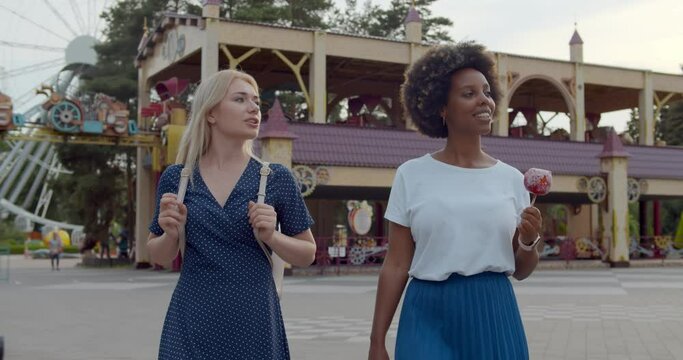 Happy Young Women Eating Candy Sweet And Walking At Amusement Park.