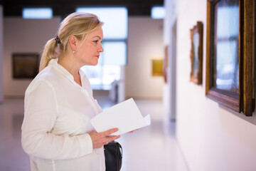 Senior female visitor holding guidebook standing at painting exhibition in art museum