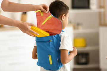 Mother putting school lunch in backpack of her little son at home