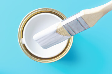 Open can of white paint and paintbrush on blue background top view.