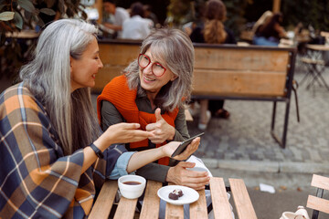 Smiling senior woman shows thumb up to friend holding cellphone at small table with coffee and...