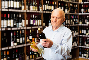 Serious elderly man chooses between red and white wine in a liquor store.
