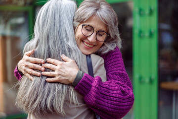 Happy senior woman with glasses in knitted jacket hugs best friend with long silver hair meeting on...