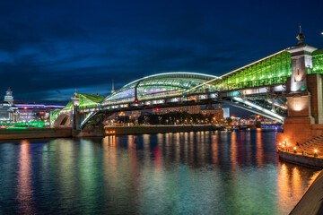Obraz premium View of the colorful Bogdan Khmelnitsky bridge illuminated at night reflecting in the Moskova river. Moscow Kiyevsky railway station at night. Moscow, Russia