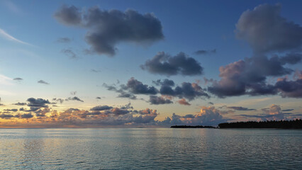 Naklejka premium Sunrise in the Maldives. There are picturesque blue and pink clouds in the azure morning sky. Reflection and ripples on the ocean surface. An island is visible on the horizon