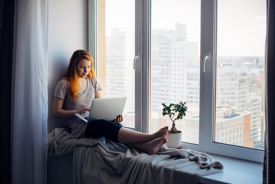 Beautiful positive girl using laptop, sitting on the windowsill in city apartment. Young red-haired woman working at home. Freelance concept.