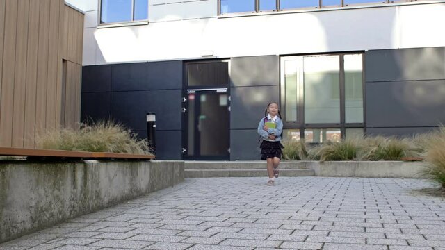 An elementary school student rushes home after school. A little girl with pigtails and a backpack runs against the background of the school building.