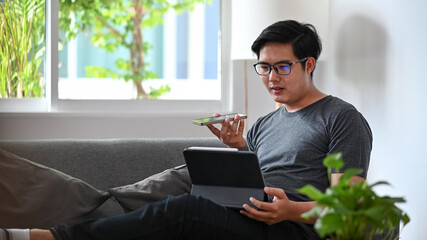 Young man talking on speaker phone and working with computer tablet on sofa.
