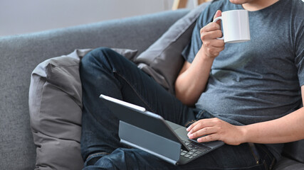 Cropped shot casual man drinking coffee and using computer tablet on couch.