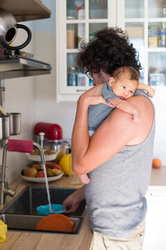 Father holding newborn while cooking in kitchen. Profile portrait, vertical shot.