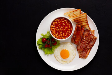 Traditional Full English breakfast, on a black background, fried eggs with fried bacon, sausages, white beans ,cherry tomatoes, horizontal, no people,