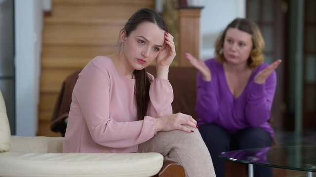Overwhelmed Caucasian young woman sitting at home as angry mother shouting gesturing at background. Portrait of sad stressed beautiful millennial arguing with parent indoors. Family conflict concept