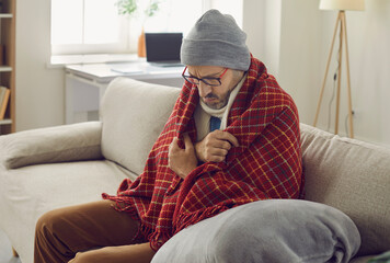 It's winter and it's cold in the house. Man having problems with central heating in wintertime. Dissatisfied man wearing hat and all wrapped in warm wool blankets sitting on sofa at home and freezing