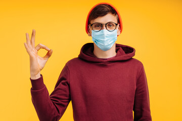 man in protective medical mask shows ok sign, standing against yellow background, in orange hat and red sweatshirt