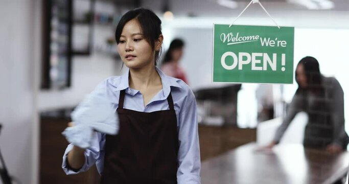 Chinese look style young woman coffee shop owner using clothe to wipe and clean clear glass wall at the front door of cafe before open for serve customer.
