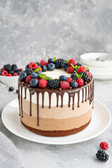Three chocolate mousse cake with chocolate glaze, blueberries, raspberries and blackberries on top on a white plate on a gray concrete background. Cake with three layers of different taste. Copy space
