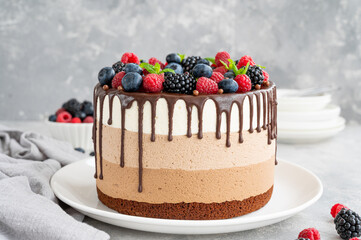 Three chocolate mousse cake with chocolate glaze, blueberries, raspberries and blackberries on top on a white plate on a gray concrete background. Cake with three layers of different taste. Copy space