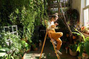 Smiling woman gardener in orange overalls sitting on stepladder in green house, resting, takes a...