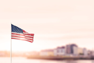 Waving flag of United States of America in focus. Modern town out of focus in the background. Warm sunny day. Sun flare. Abstract city concept