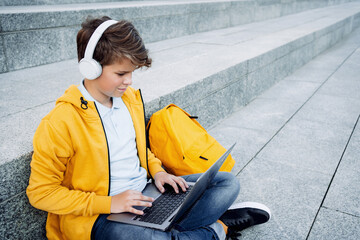 Child caucasian teen boy in yellow hoodie with headphones sitting outdoors using laptop. Remote or distant learning on the go. Blogging surfing. Homeschooling. Lockdown. Video call. Copy space.