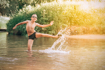Cheerful funny boy 7-10 jump in river wearing swimming goggles, splashing in rays of sun